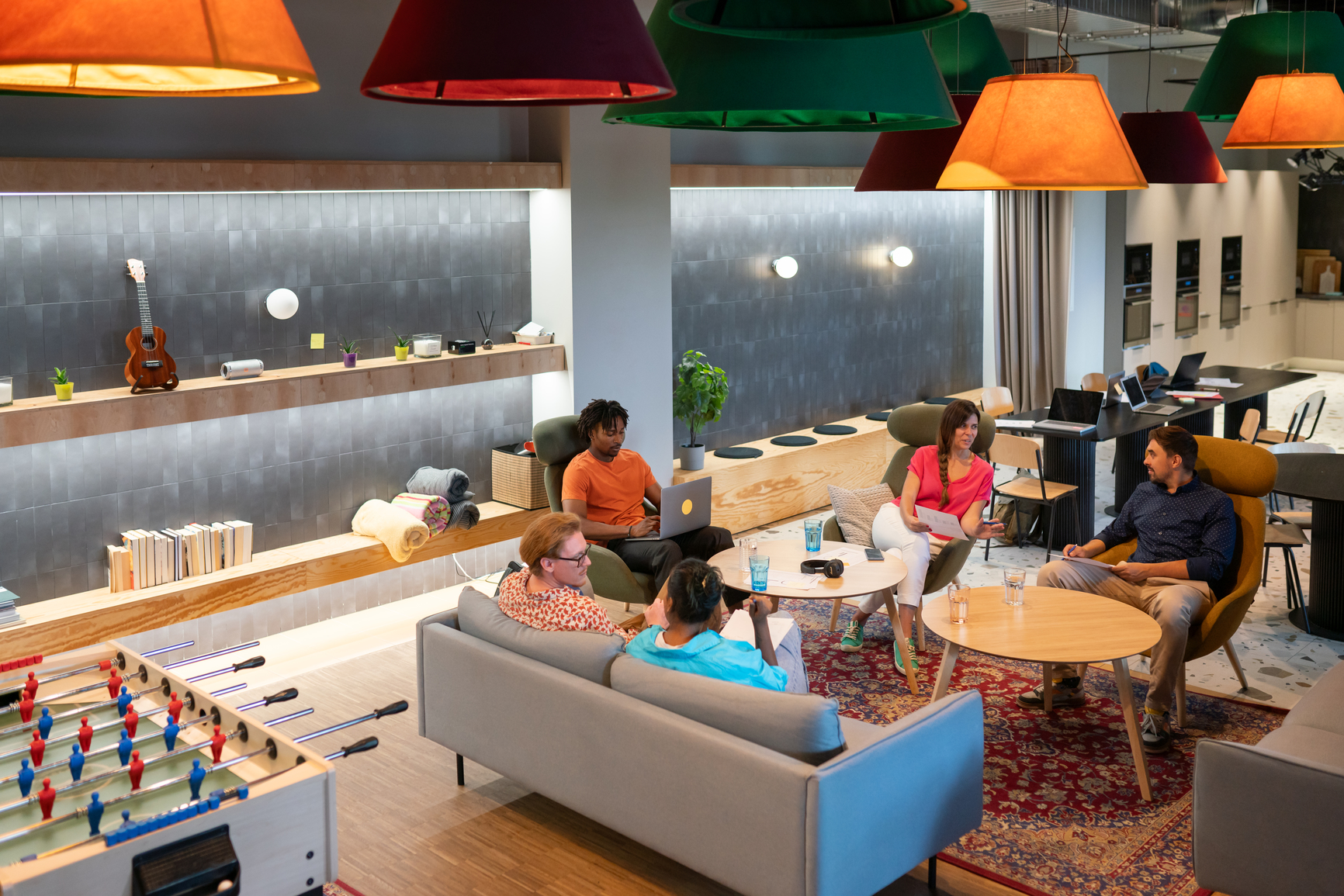 Informal meeting of colleagues at co-working lounge area. Group of multiracial people having a conversation. They are sitting on the sofa and armchairs in a round. The place looks modern, illuminated by stylish design colourful lamps and led illumination.